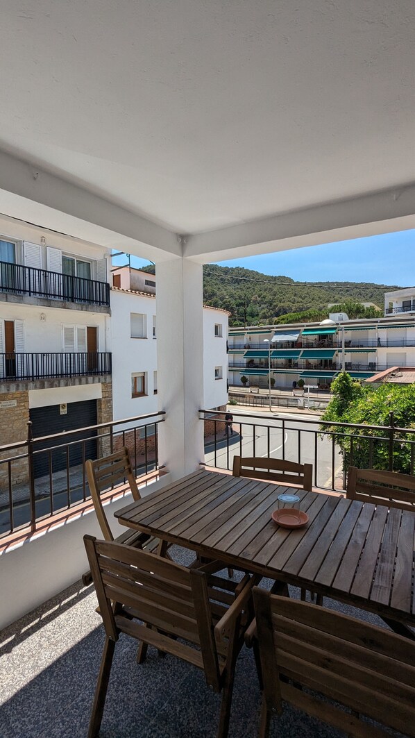 Un balcón con una mesa y sillas de madera, con vistas a una zona residencial con edificios y un paisaje montañoso.