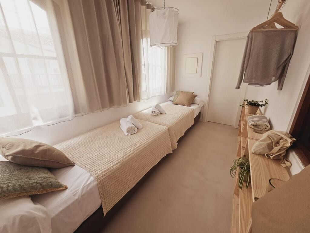 A hotel room with two single beds, a hanging light fixture, a small table with a plant, and a coat rack.