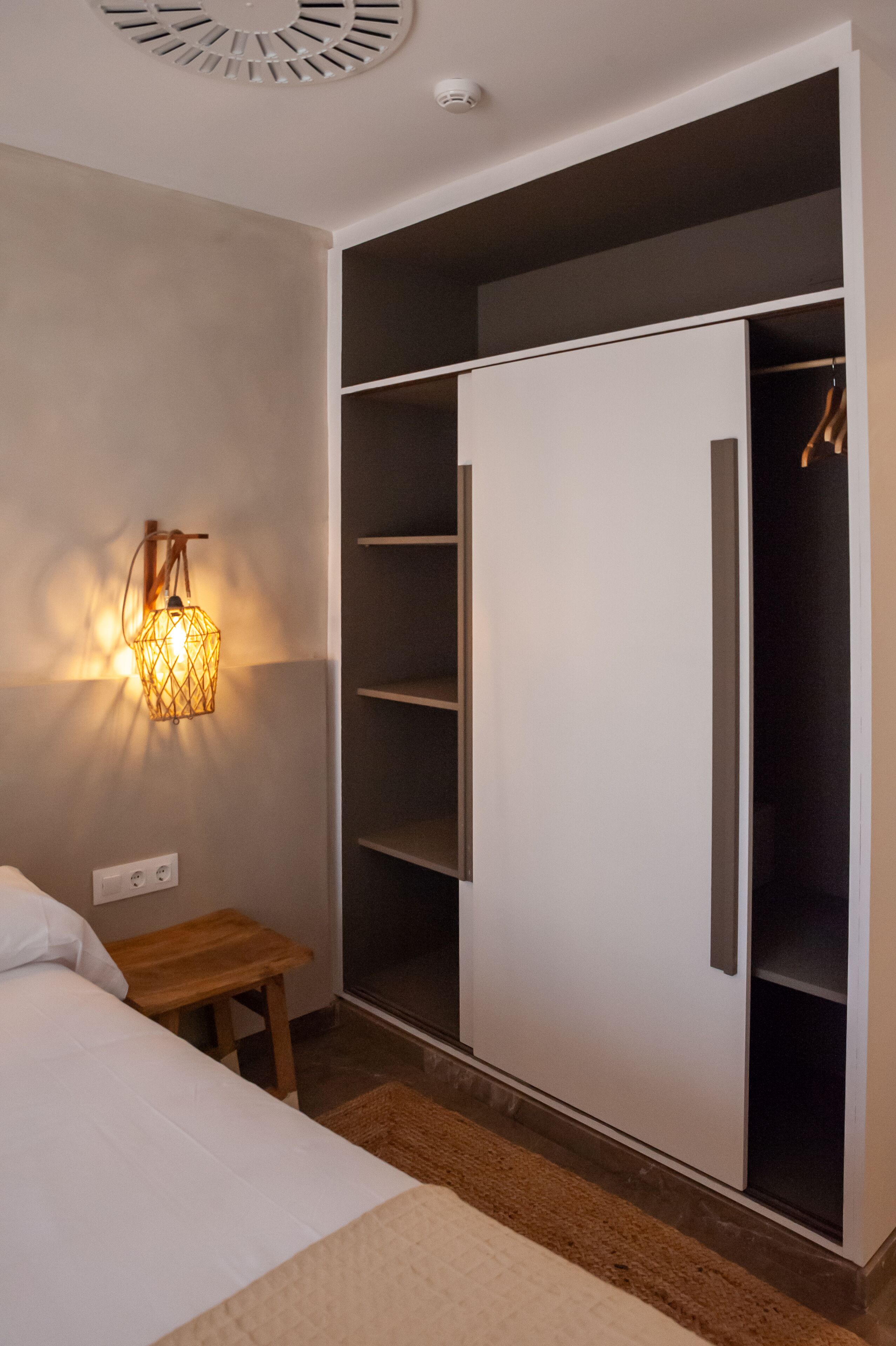 A bedroom with a bed, a bedside table, a wardrobe, and a wall-mounted light fixture.