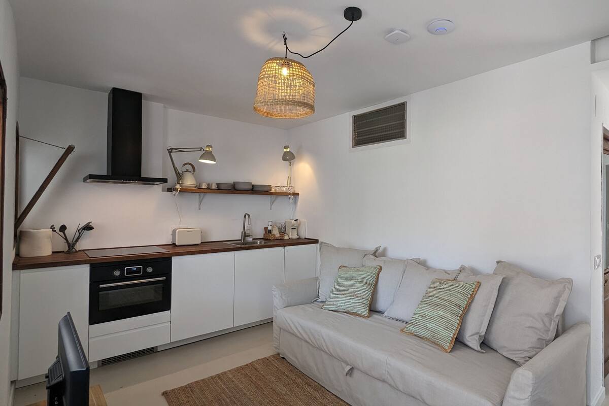 Una cocina moderna con armarios blancos, horno integrado y encimera de madera. En la zona de estar, hay un sofá color beige con almohadones a rayas.