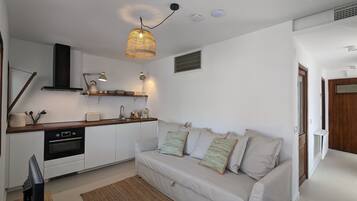 Una cocina moderna con armarios blancos, horno integrado y encimera de madera. En la zona de estar, hay un sofá color beige con almohadones a rayas.