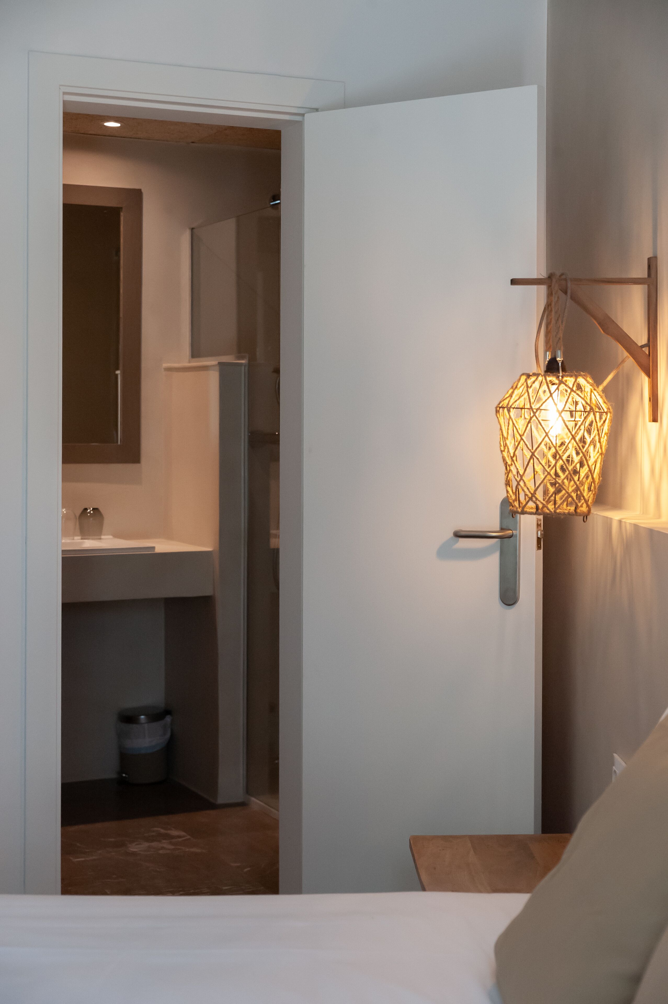 A hotel room with a bed, a bedside table, a nightstand, a wall-mounted lamp, a door, and an open closet leading to a bathroom with a sink and a mirror.