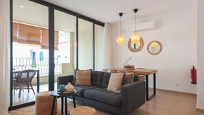 A modern living room with a sofa, a small table, and a dining area. There are two pendant lights, a fire extinguisher, and a large window with blinds. - Hostal Es Niu de Tamariu (Palafrugell)