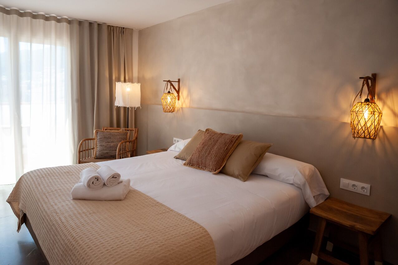 Cama bem arrumada com manta bege e almofadas, abajur e mesinha de cabeceira de madeira.