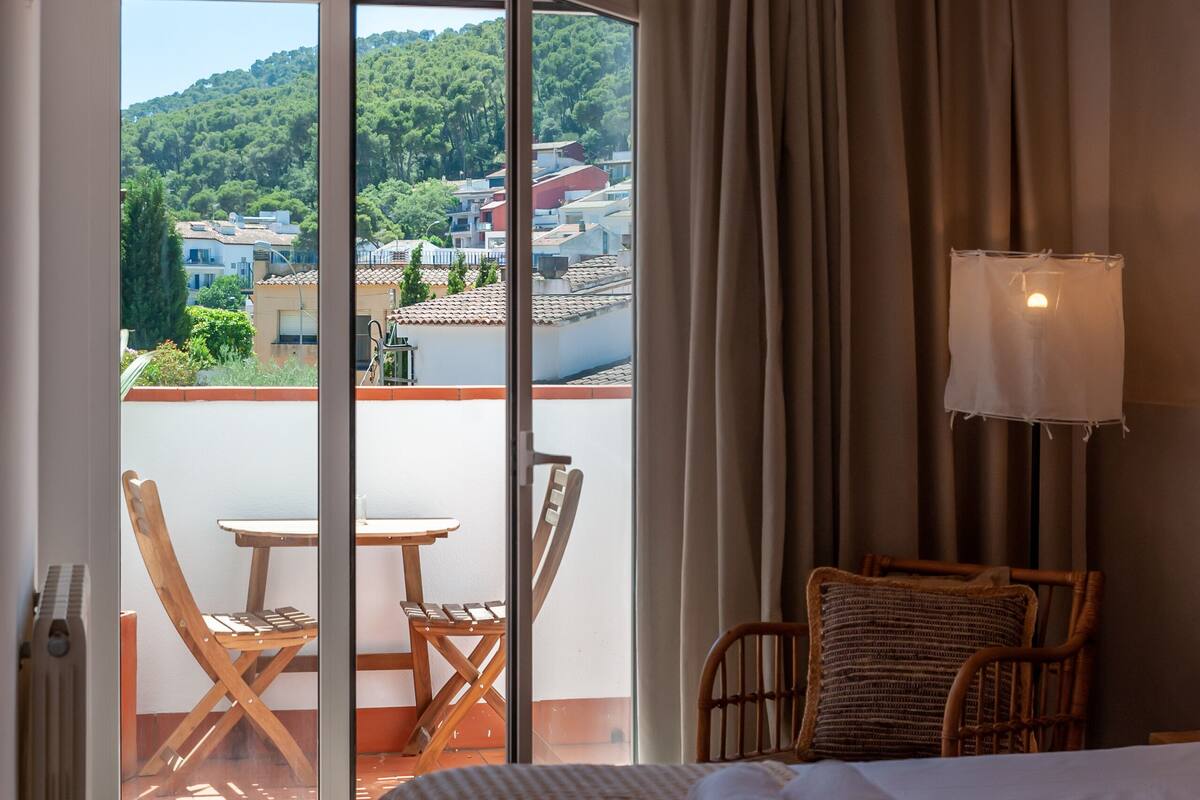 Habitación de hotel con cama, silla, mesa, vistas a un balcón y una lámpara.