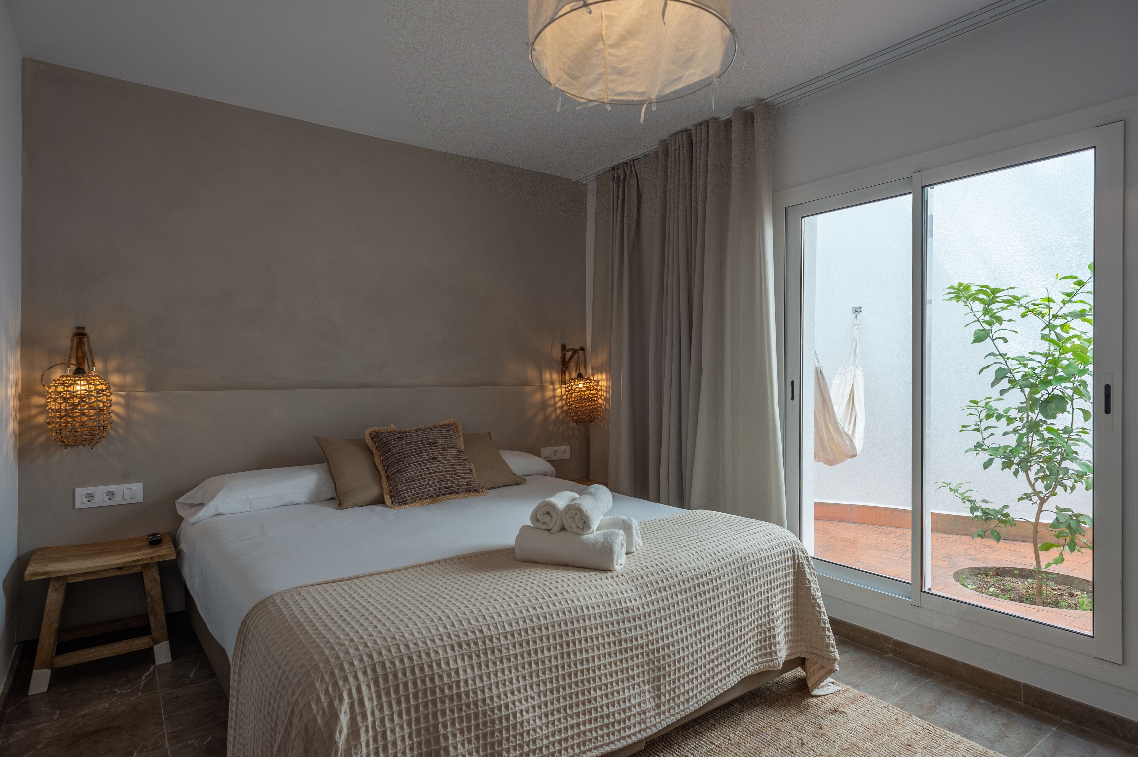 A bedroom with a large bed, two wall-mounted lamps, a small bedside table, and a view of the outdoors through a sliding glass door.