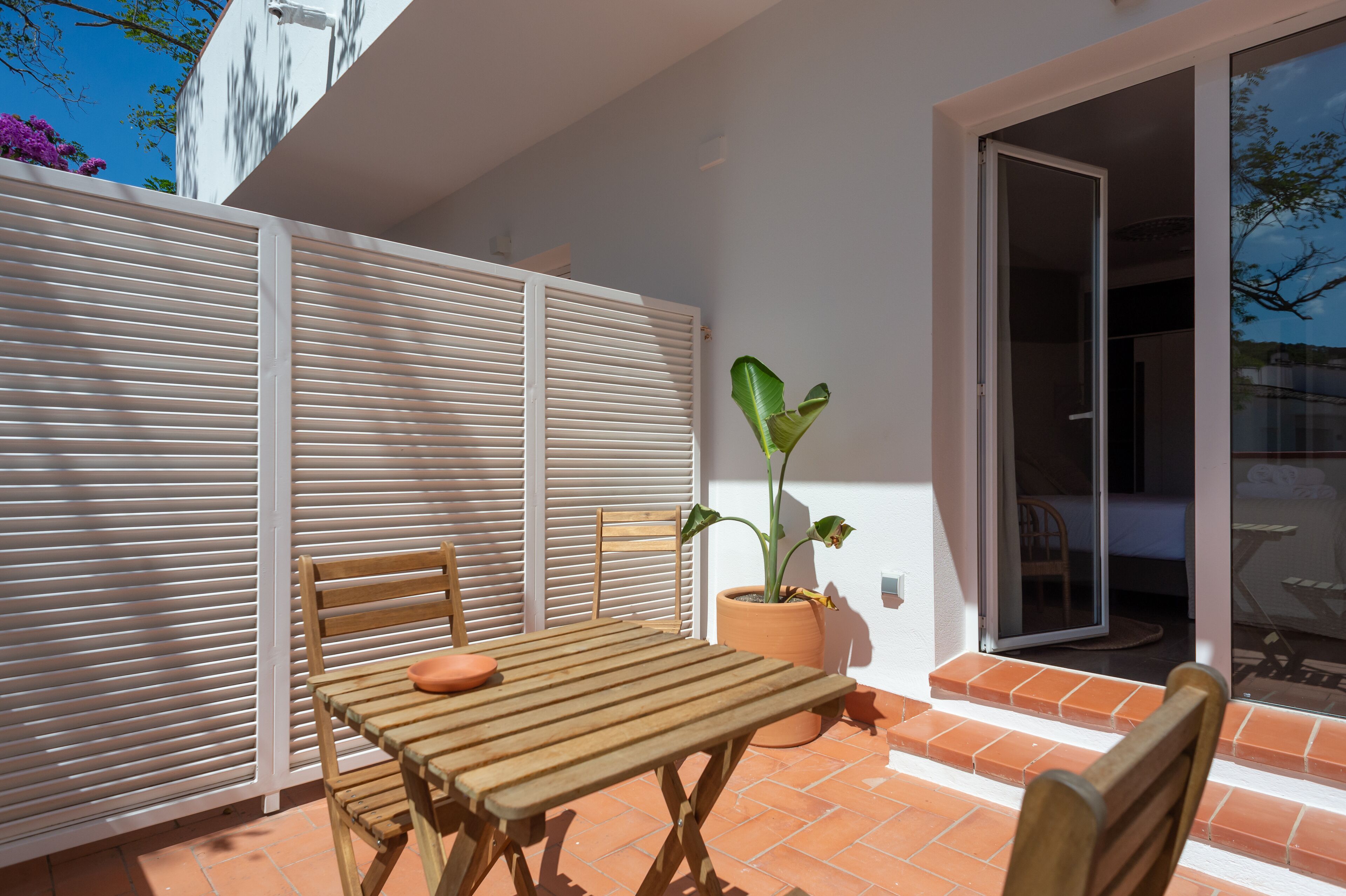 An outdoor patio with a wooden table and chairs, a potted plant, and a sliding glass door leading to an interior room.