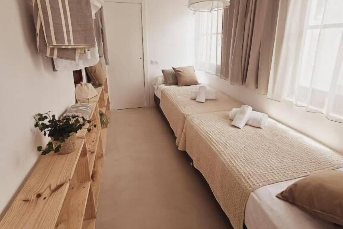 Una habitación de hotel pequeña y alargada, con un banco de madera largo, dos camas, una lámpara colgante y un ventanal con cortinas.
