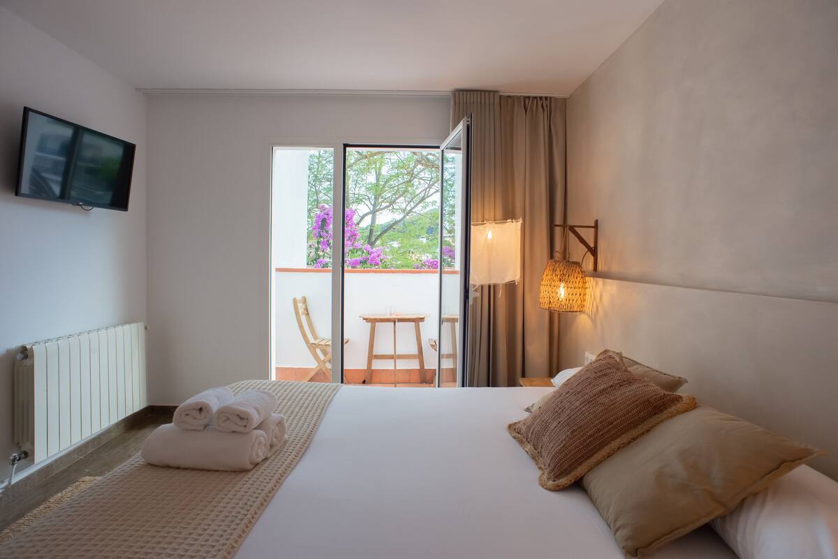 Habitación de hotel con cama, televisor montado en la pared, balcón con mesa y sillas, y vistas a árboles y flores.
