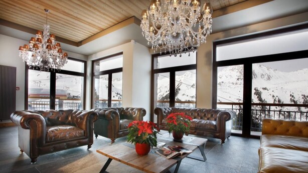 A spacious lounge with leather sofas, a wooden coffee table, and a large chandelier. Large windows offer views of a snowy mountain. - Wifi, 50m from resort center, fitness, swimming pool, sauna, hammam, balcony, tv, ski locker, 45m² (Les Arcs)