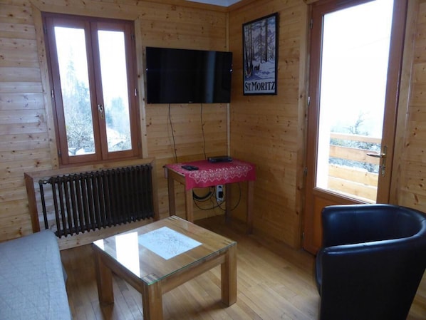 A wooden cabin interior with a glass table, a red tablecloth, a black chair, a mounted TV, and a poster on the wall.