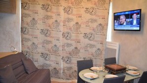 A room with a patterned wallpaper, a sofa, a dining table set with plates and glasses, and a television mounted on the wall. - 10m from ski slopes, 13rd floor, view mountain, tv, 25m², Les Menuires (Les Menuires)