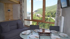 A dining area with a table set for four, a sofa, and a large window offering a view of a mountainous landscape. - 10m from ski slopes, 13rd floor, view mountain, tv, 25m², Les Menuires (Les Menuires)