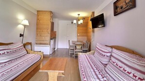 Een knus vertrek met twee gestreepte banken, een houten salontafel, een eettafel met stoelen, een klein keukentje en een aan de muur bevestigde televisie.
