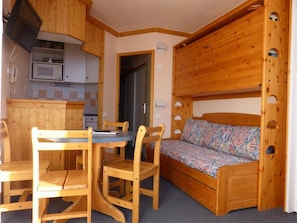 Une pièce compacte comprenant un lit superposé en bois, une petite kitchenette, une table à manger avec des chaises et un lit simple avec un matelas à motifs.