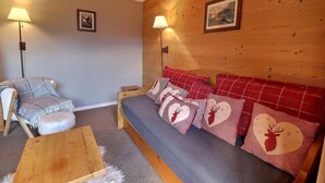 Una habitación acogedora con una pared de madera, un sofá gris con cojines blancos y rojos a cuadros, una silla de madera y dos lámparas de pie.