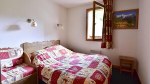 A bedroom with a wooden bed, a bedside table, a wall-mounted lamp, a window with curtains, and a painting on the wall. - 150m from ski slopes, 2nd floor, balcony, tv, ski locker, 45m², Valloire (Valloire)