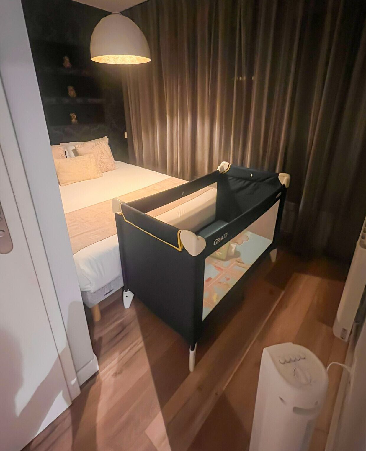 Une chambre d’hôtel avec un lit, un berceau noir et blanc, une grande fenêtre avec des rideaux et un plancher en bois.