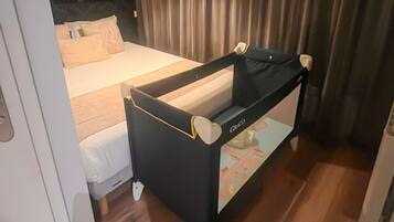 Une chambre d’hôtel avec un lit, un berceau noir et blanc, une grande fenêtre avec des rideaux et un plancher en bois.