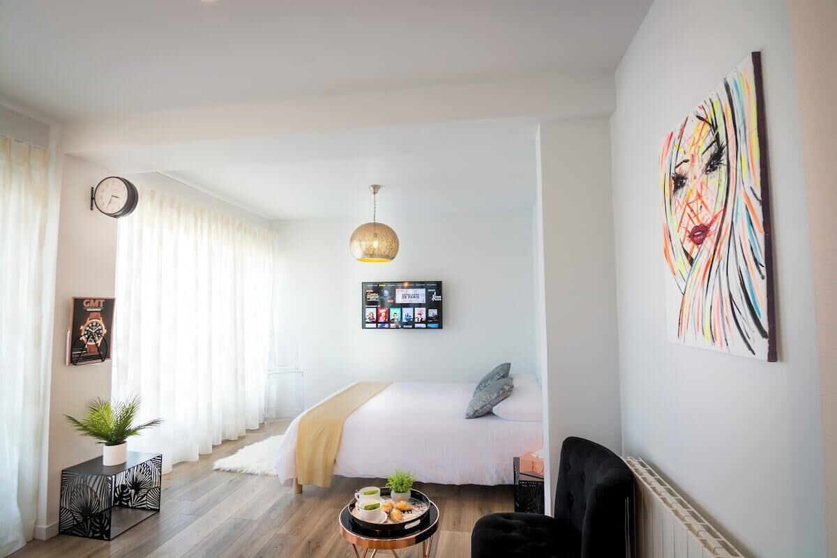 A modern hotel room with a bed, a round table with a tray of food, a black armchair, a hanging light fixture, and a wall art piece.