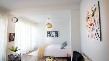 A modern hotel room with a bed, a round table with a tray of food, a black armchair, a hanging light fixture, and a wall art piece.