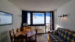 A room with a wooden dining table and chairs, a plaid sofa, a window with a view of a snowy landscape, and a framed picture on the wall.