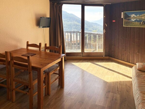 A room with a wooden dining table, chairs, a sofa, a TV, and a large window with a mountain view.