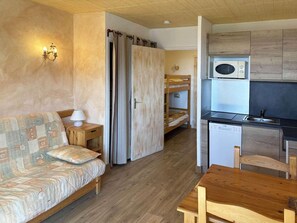 A compact room with a sofa, a small table, a kitchenette with a microwave and a dishwasher, and a wooden ceiling.
