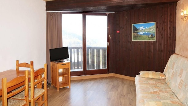 Een kamer met een houten vloer, een houten tv-meubel met een flatscreen televisie, een houten eettafel met stoelen en een gestreept bankje.