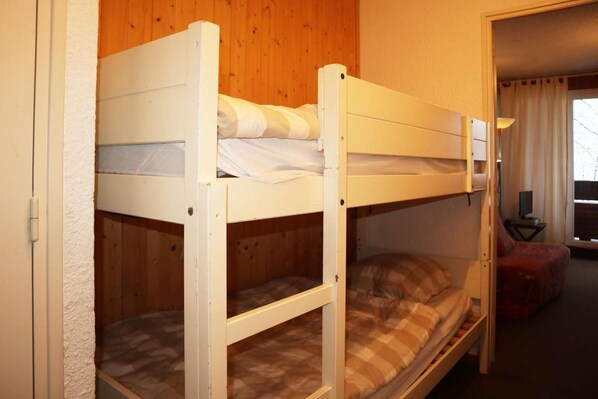 A bunk bed room with a wooden floor, a small table with a lamp, and a window with curtains.