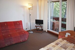 A room with a red sofa, a television on a small table, a floor lamp, and a view of the outdoors through a large window.