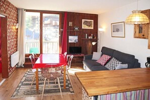 A living room with a wooden table, a sofa, and a TV. There is a window with curtains and a view of buildings outside. - 15m from ski slopes, 2nd floor, view ski slopes, balcony, 40m², Les Orres (Les Orres)