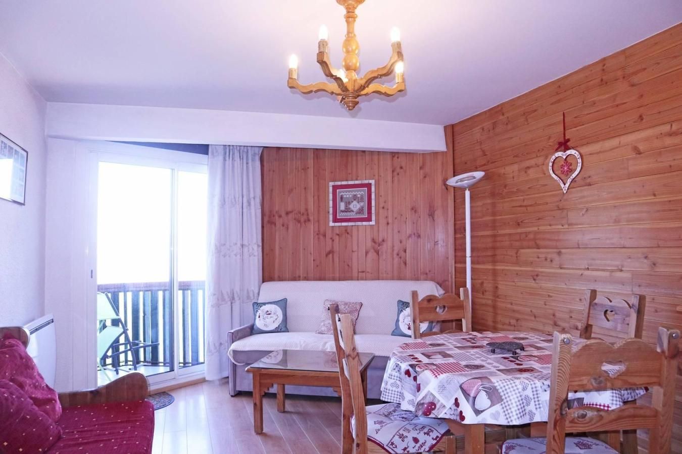 A cozy room with a wooden wall, a chandelier, a sofa, a dining table with chairs, and a large window with a view.