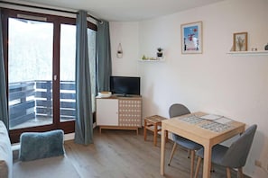 A furnished room with a balcony, a television, a dining table with chairs, and a small cabinet.
