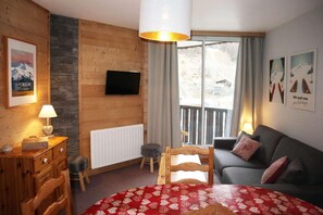 A cozy room with a wooden wall, a sofa, a red patterned bedspread, a television, and a window with a view.