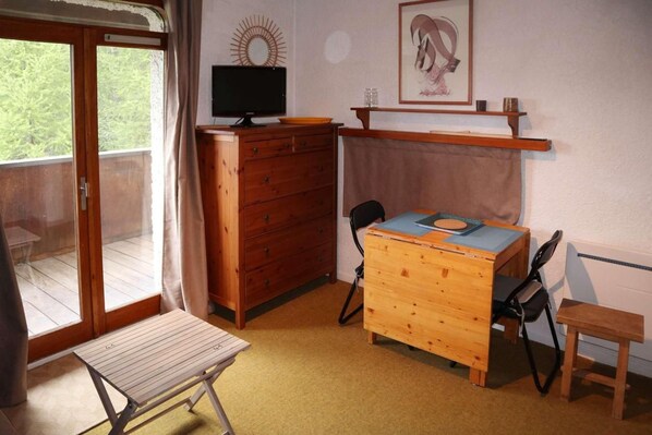 Une pièce avec un bureau en bois, une chaise, un téléviseur posé sur une commode et une fenêtre donnant sur un balcon.