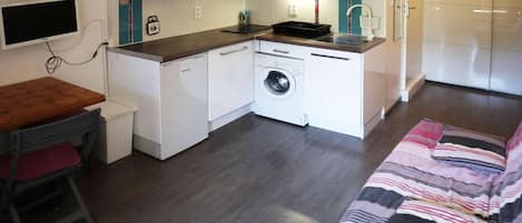 Cocina compacta equipada con microondas, horno y lavadora. Hay una pequeña mesa de comedor con una silla y una cama con manta a rayas.