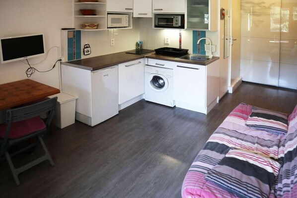 A compact kitchenette with a microwave, oven, and washing machine. There is a small dining table with a chair and a bed with a striped blanket.