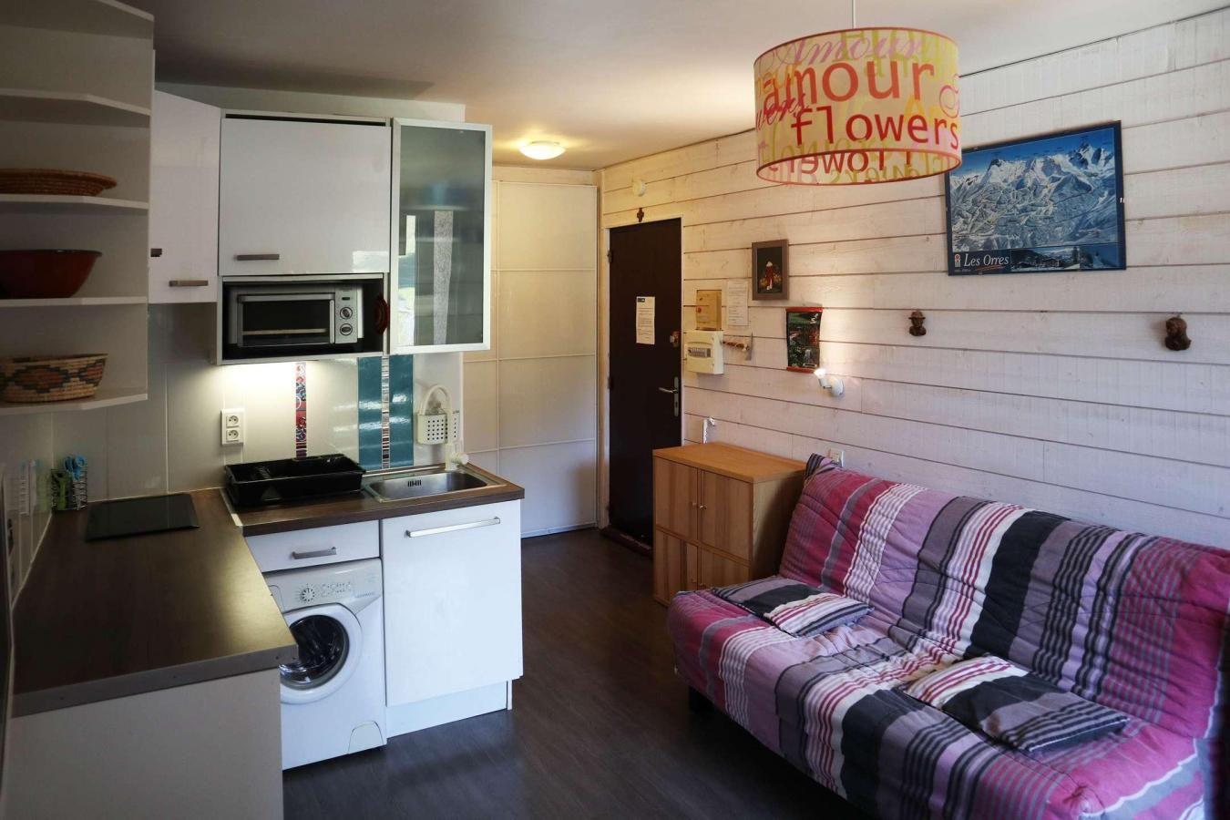 Una cocina compacta con microondas, lavaplatos y lavadora. Hay un sofá con una manta de colores y una lámpara de pared.