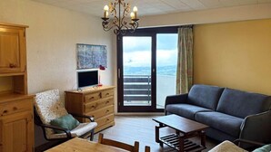 A living room with a wooden dining table, a sofa, a chair, and a TV. There is a balcony with a view of the city.
