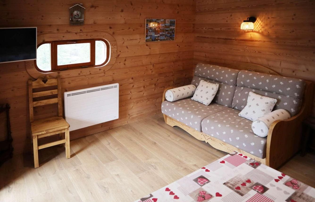A cozy room with a wooden floor, a sofa with patterned cushions, a small wooden chair, a radiator, and a wall-mounted light fixture.