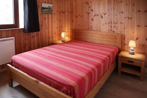 Una habitación con paneles de madera, una cama, dos mesitas de noche con lámparas, un radiador y una ventana con vista a una montaña.