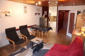 Een gezellige woonkamer met een rode bank, twee zwarte fauteuils, een klein tafeltje en een keuken op de achtergrond.