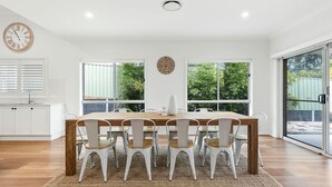 A dining area with a wooden table and white chairs, large windows with a view of greenery, a wall clock, and a white cabinet with a sink. - Gone Coastal at Narrawallee (Narrawallee)