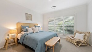 A bedroom with a bed, bedside tables, a chair, and a window with blinds. - Gone Coastal at Narrawallee (Narrawallee)