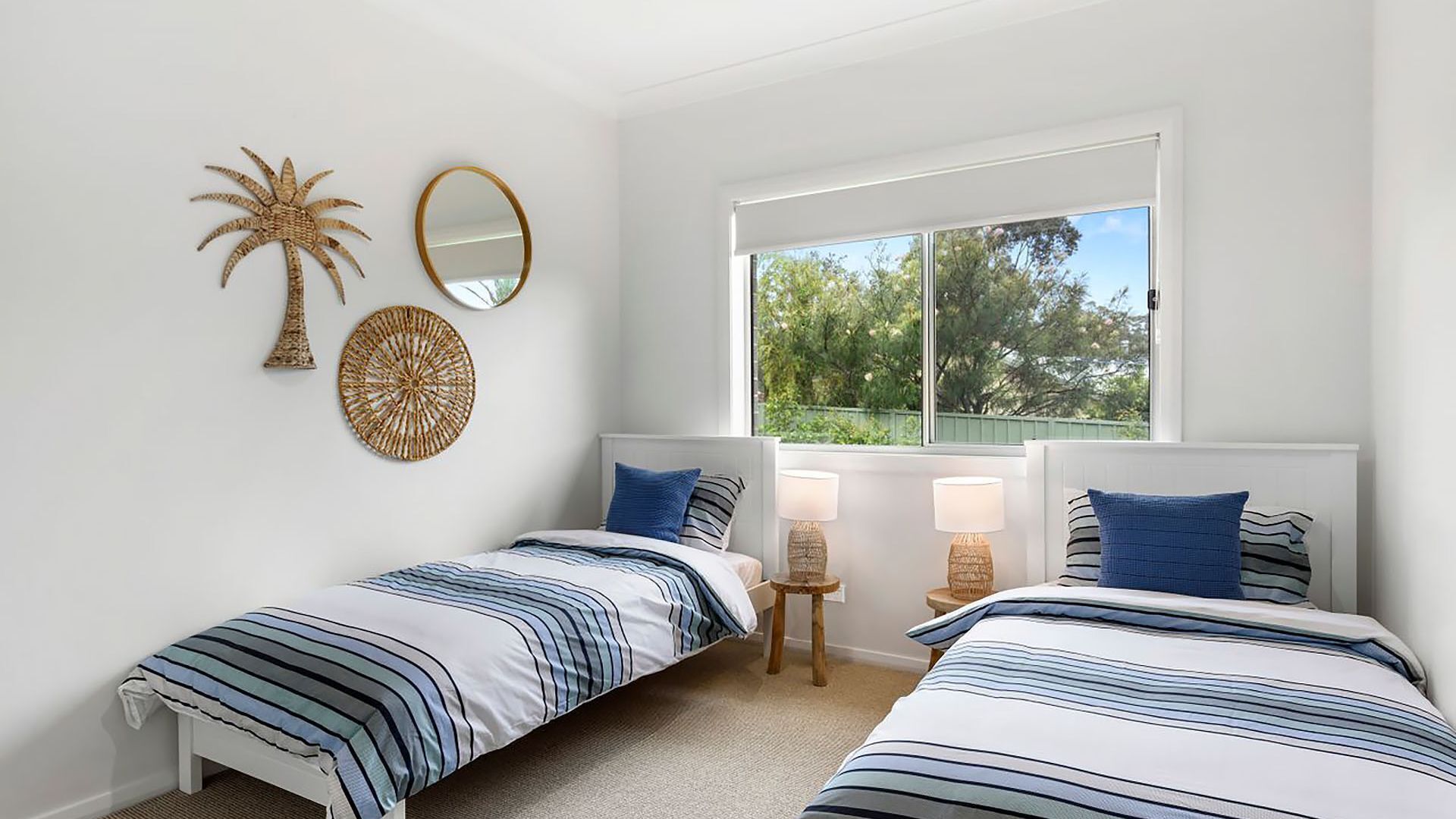 A bedroom with two beds, a window with a view of greenery, and wall decor including a palm tree and a circular woven piece.