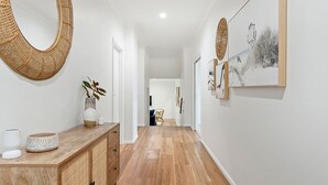A hallway with a wooden floor, a woven wall hanging, a vase with a plant, and framed artwork on the wall. - Gone Coastal at Narrawallee (Narrawallee)
