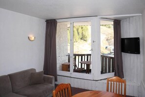 A room with a sofa, a table, two chairs, a television mounted on the wall, and a sliding glass door leading to an outdoor area with a wooden deck and a view of trees. - 15m from ski slopes, 5th floor, view resort, balcony, 28m², Les Orres (Les Orres)