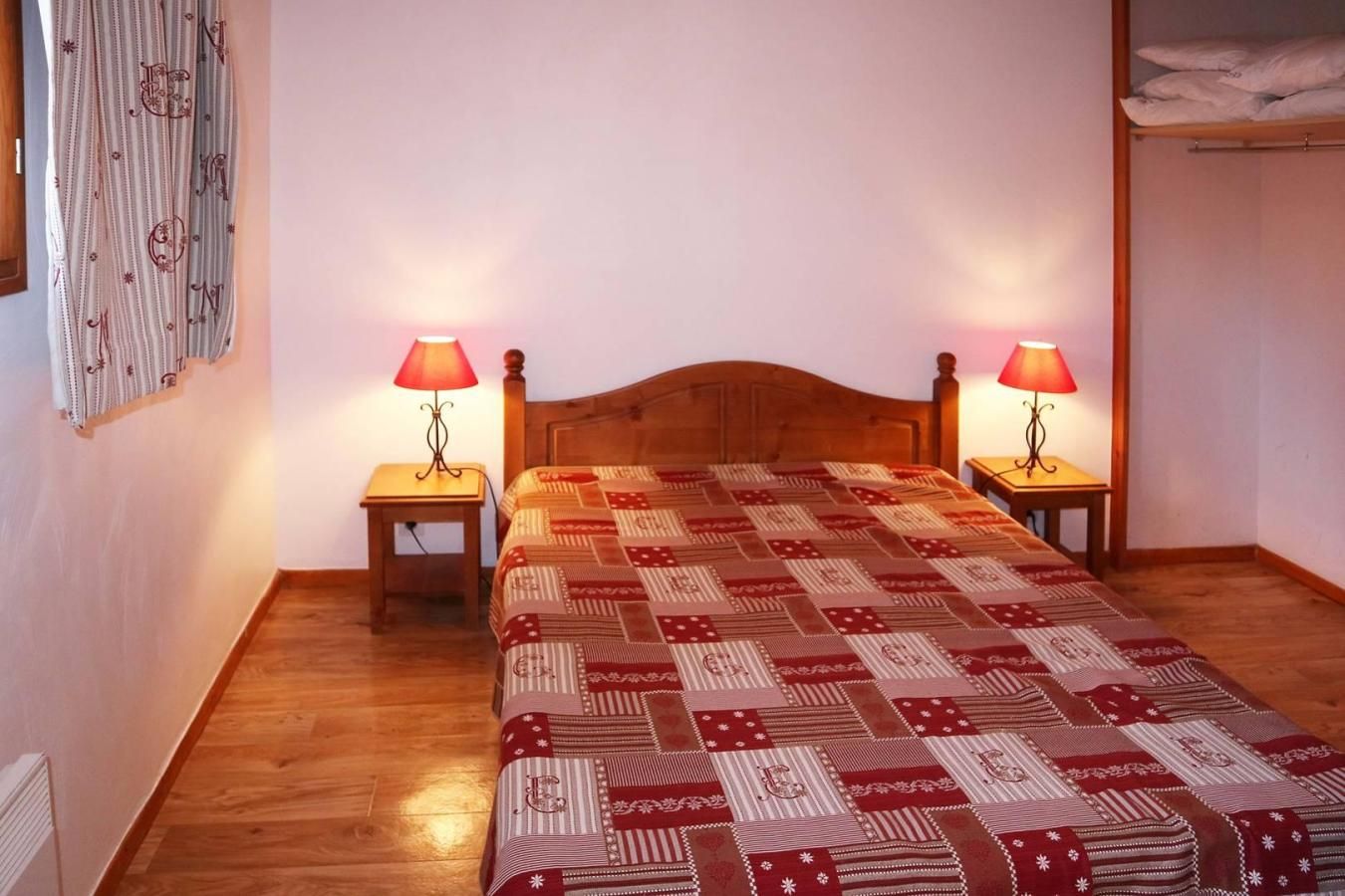 A bedroom with a wooden bed, two bedside tables with lamps, a patterned bedspread, and a wall with a curtain.