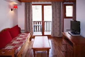 A room with a wooden sofa, a wooden table, a television on a wooden stand, and a sliding glass door leading to an outdoor area.